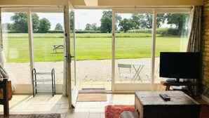 En stue med fjernsyn, sofa og bord. En skydedør fører ud til en terrasse med græsplæne og bord.