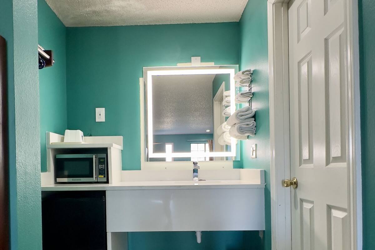 Une salle de bain avec des murs de couleur turquoise, un lavabo blanc, un miroir avec éclairage intégré, un four à micro-ondes et une poubelle.