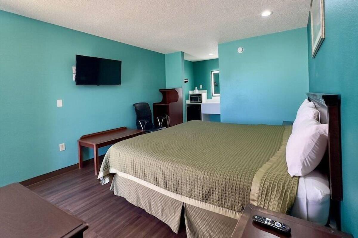 Une chambre d’hôtel aux murs de couleur turquoise, un lit recouvert d’une couverture verte, un bureau avec une chaise, un téléviseur fixé au mur et un four à micro-ondes.
