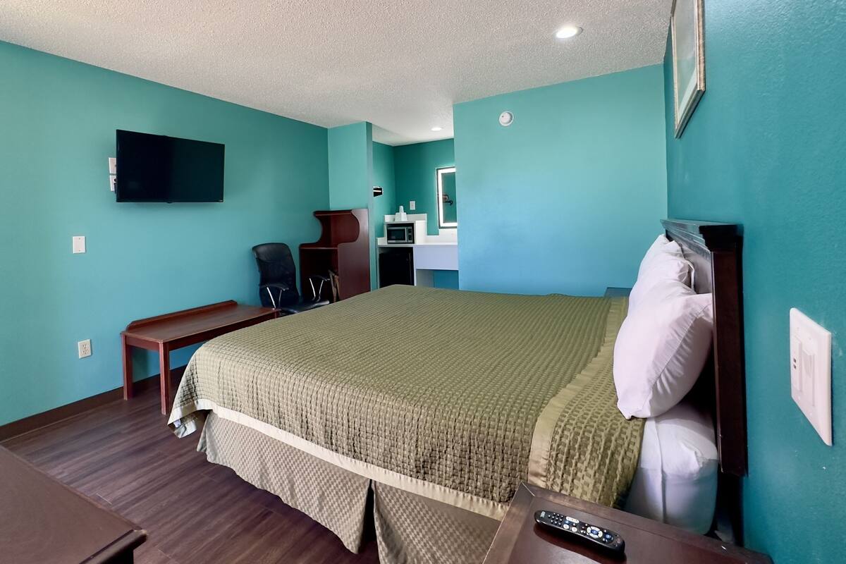 Une chambre d’hôtel aux murs de couleur turquoise, un lit recouvert d’une courtepointe à carreaux, un bureau avec une chaise, un téléviseur fixé au mur et un four à micro-ondes posé sur une petite table.