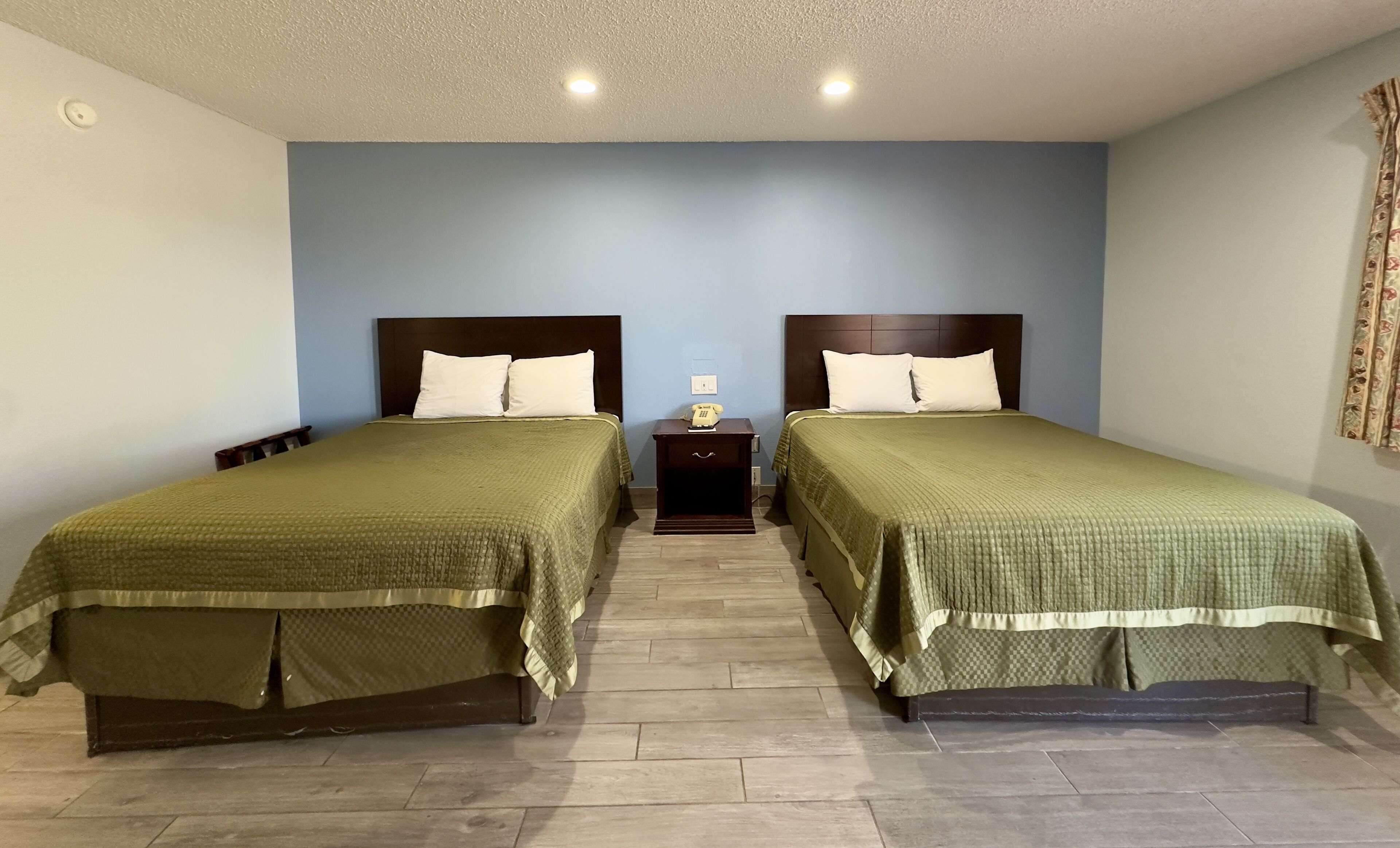 A hotel room with two single beds, a small bedside table with a lamp, and a plain blue wall.
