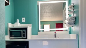 Un baño con paredes verde azulado, un lavamanos blanco, un espejo, un microondas y un cubo de basura.