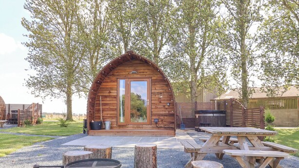 A rustic wooden cabin with a large door and windows, surrounded by a gravel area with picnic tables and a fire pit. - Mischief (Driffield)