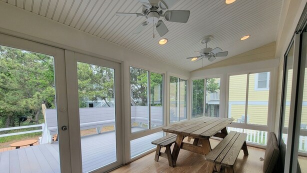 Un patio couvert avec une table et des bancs en bois, de grandes fenêtres, des ventilateurs de plafond et une vue sur le jardin arrière.