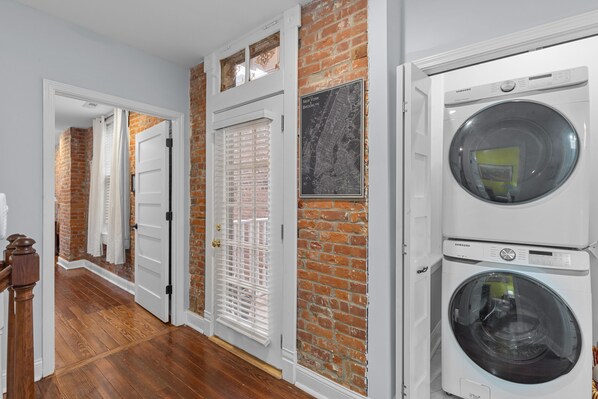 Un lavadero con dos lavadoras Samsung blancas, una pared de ladrillo y piso de madera.