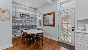 A dining area with a white table and chairs, a storage unit with shelves, and a large mirror on the wall.
