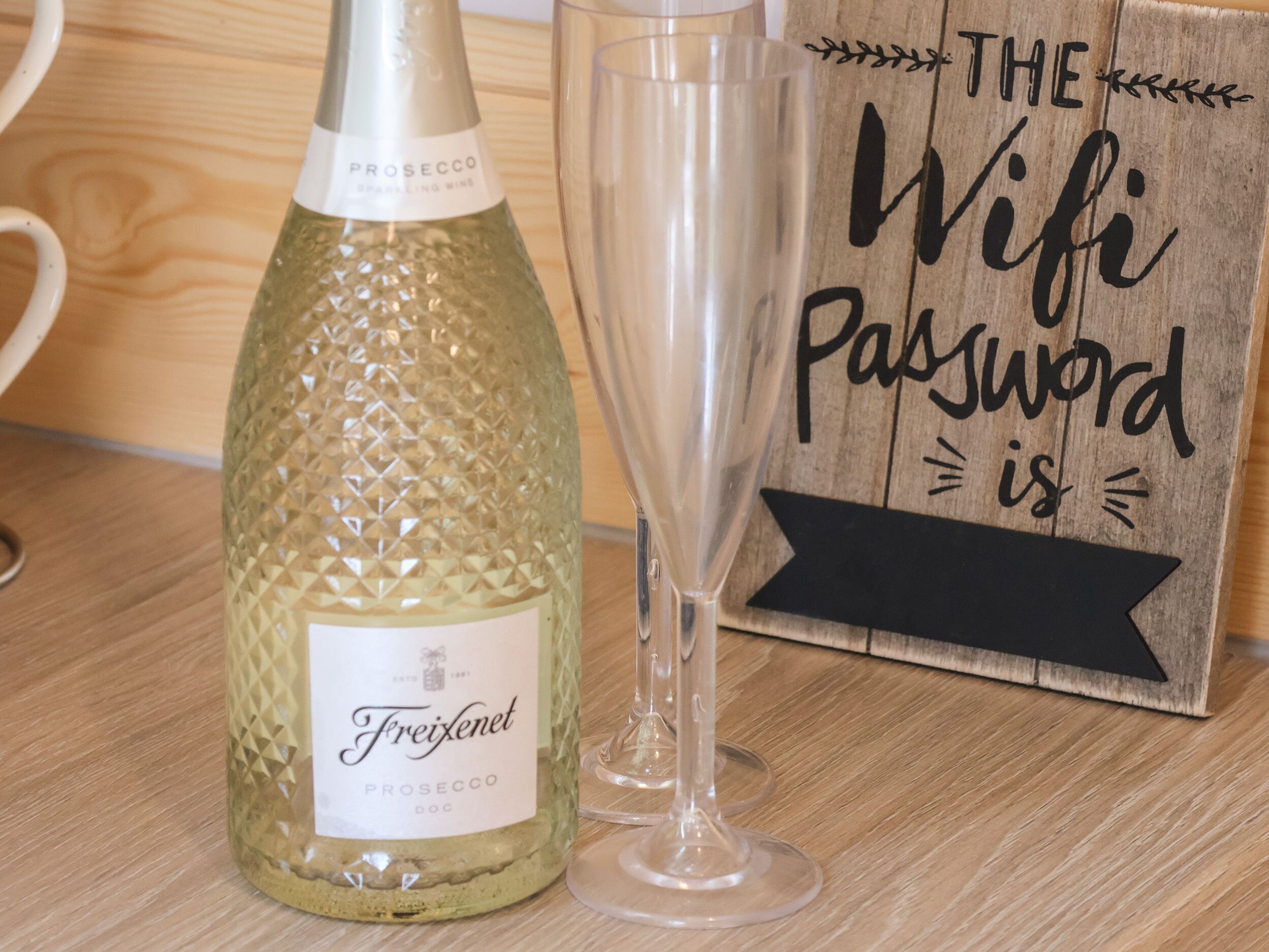 A bottle of Freixenet Prosecco DOC wine and a glass on a wooden surface with a sign that reads "The Wifi Password is".