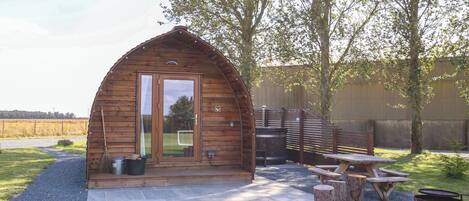 A wooden cabin with a large glass door, a patio with a table and benches, and a barbecue grill.