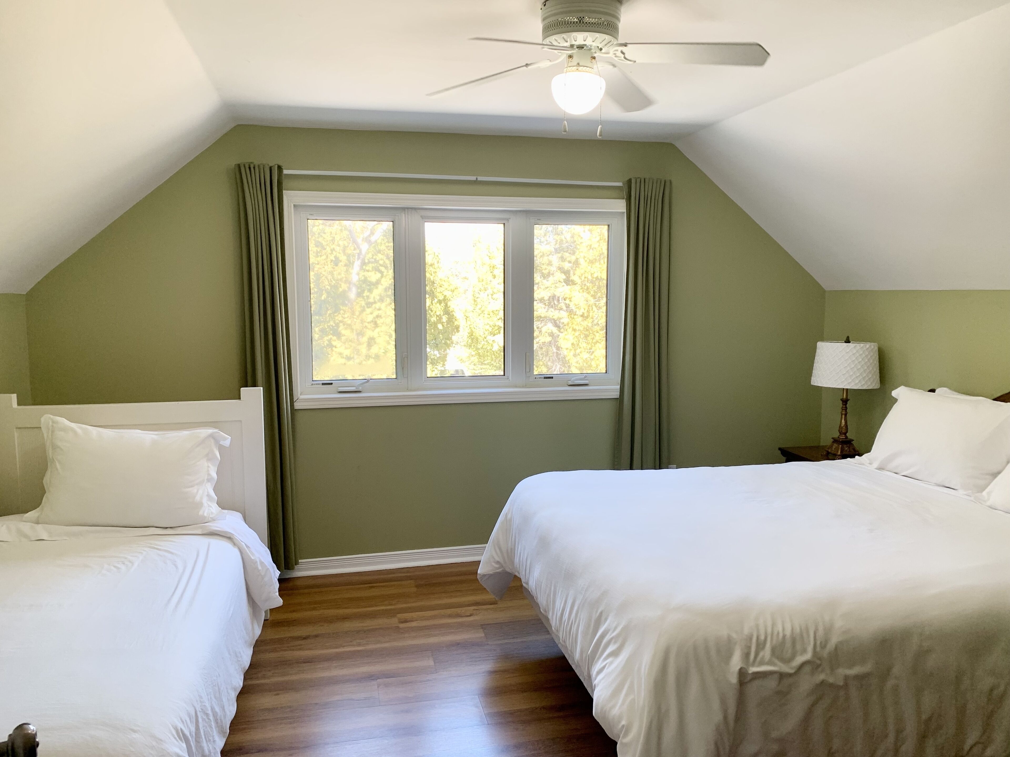 A room with two beds, a ceiling fan, a window with curtains, and a nightstand with a lamp.