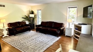 A living room with two dark brown leather sofas, a patterned rug, a white side table with a lamp, a tall floor lamp, and a television on a stand. - Cottage in the heart of Tobermory - AC & WIFI (Tobermory)