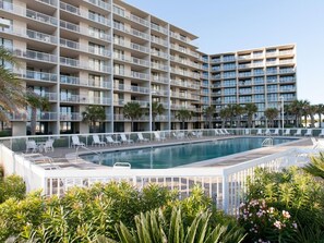 Un hotel de varios pisos con piscina rodeada de sillones de descanso y palmeras.
