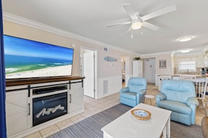 A living room with a flat-screen TV, a fireplace, two blue armchairs, a coffee table, and a ceiling fan.