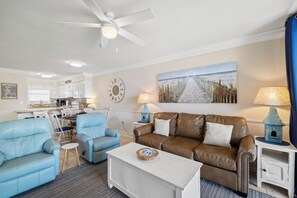 Una sala de estar con un sofá de cuero marrón, un sillón azul, una mesa de centro blanca, un cuadro de una playa y una cocina al fondo.