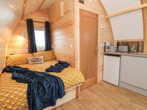 Una cabana acollidora amb interior de fusta, un llit amb roba de llit estampada, una cuineta petita i una porta que condueix a una altra habitació.