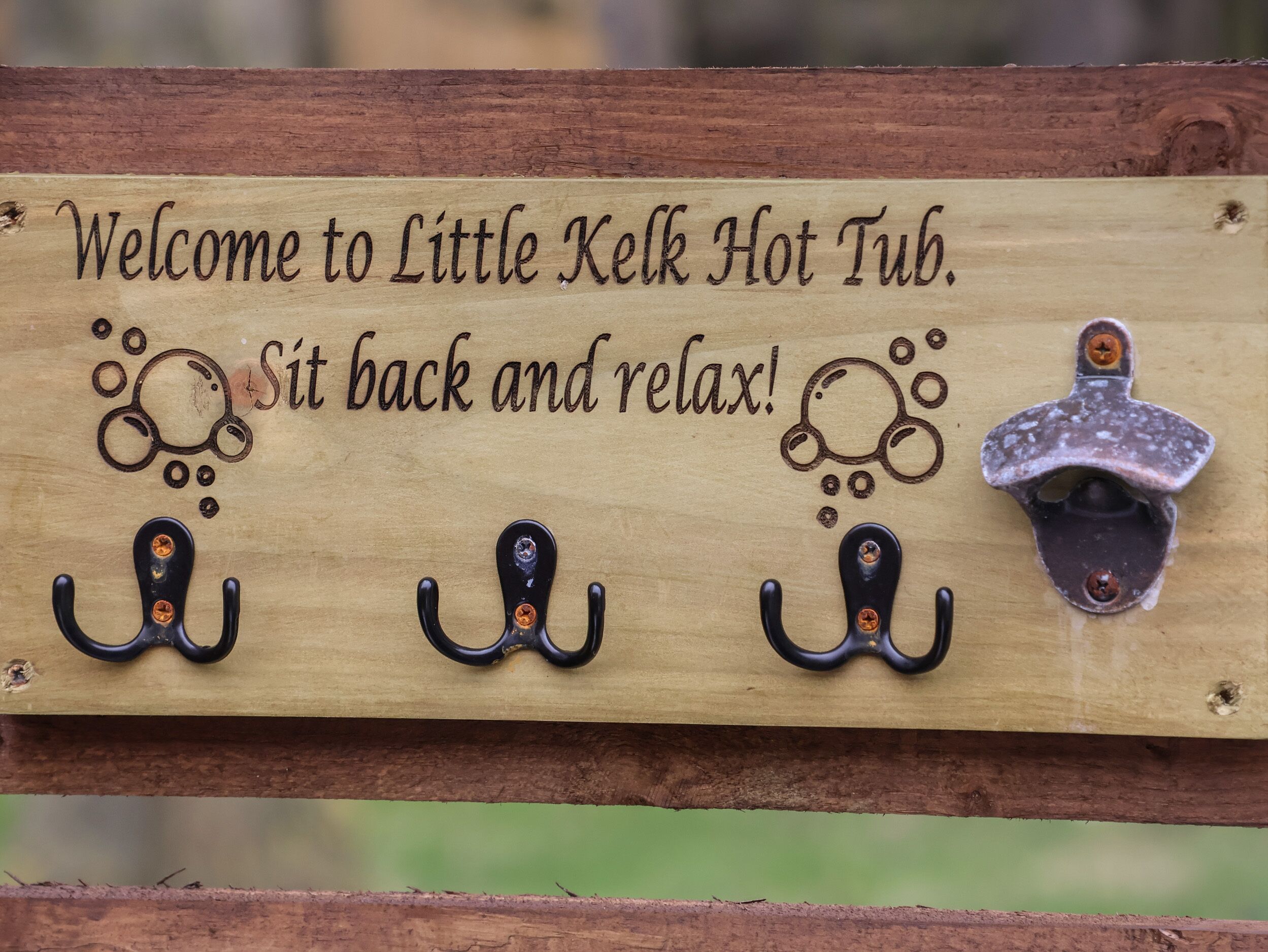 A wooden sign with the text "Welcome to Little Kelk Hot Tub. Sit back and relax!" and three black hot tub hooks.