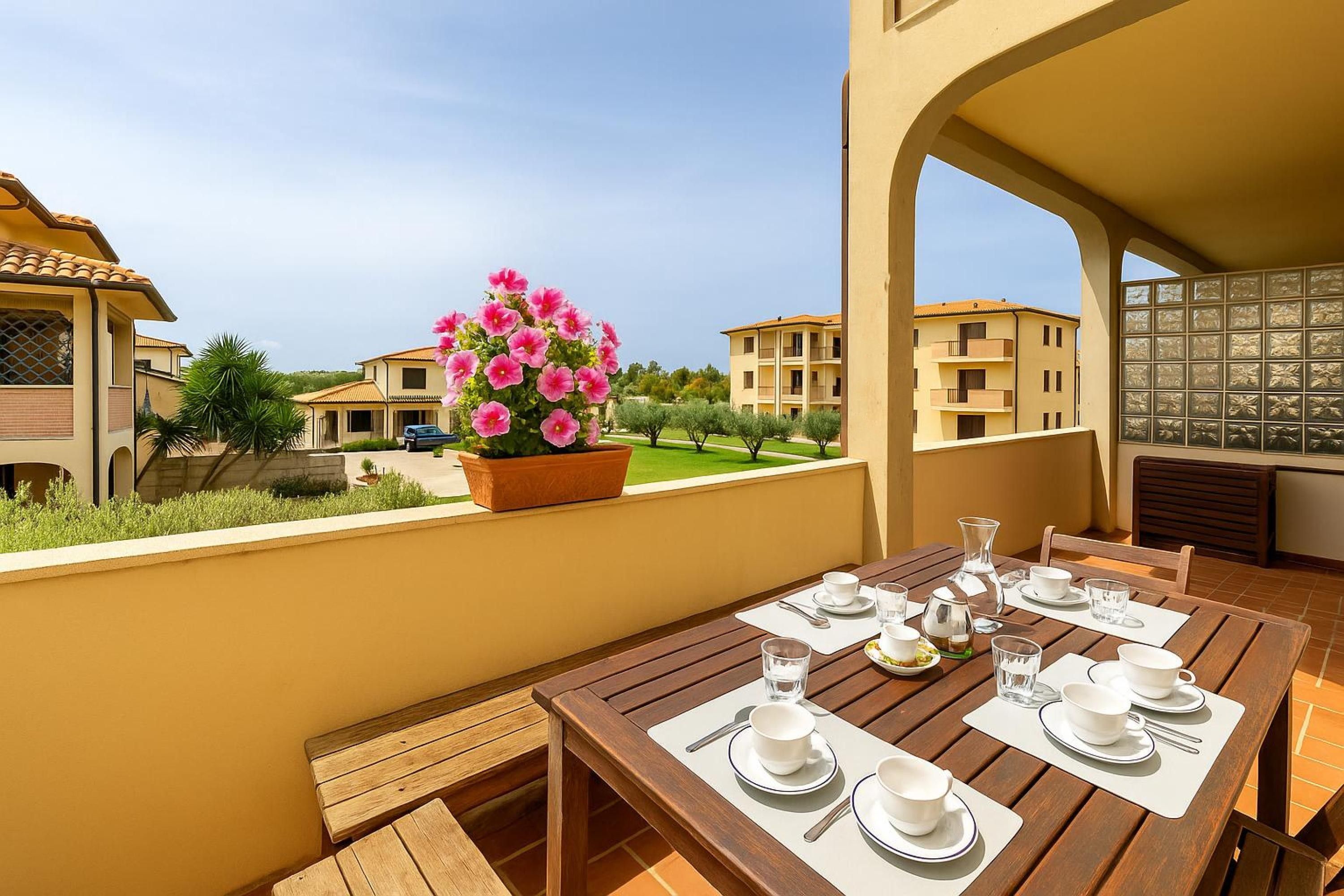A dining area with a table set for four, a potted plant with pink flowers, and a view of neighboring buildings.