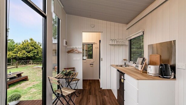 A modern kitchen with wooden flooring, a dining area with a table and chairs, and a view of the backyard through large windows. - Brumbys Run by Tiny Away (Warragul South)
