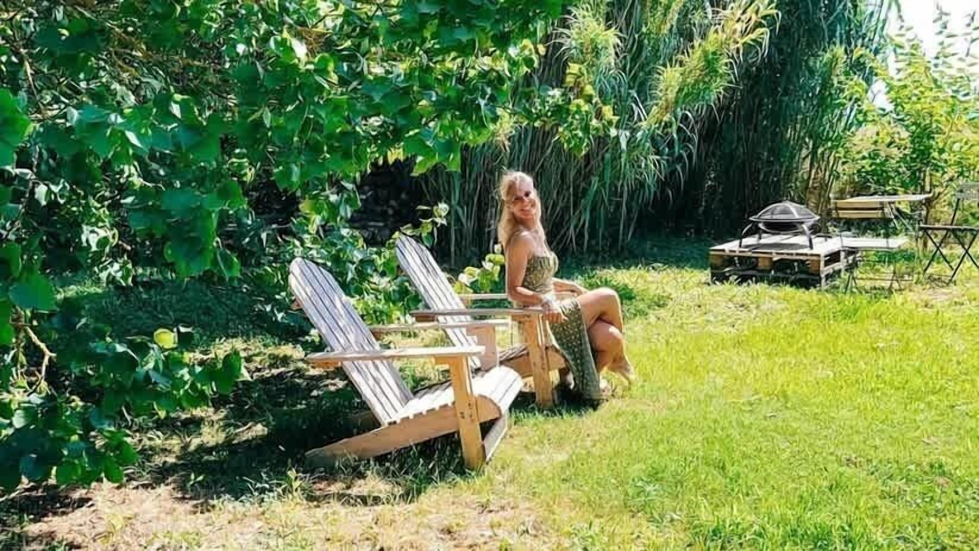 A person sitting on a wooden chair in a garden with a table and chairs in the background.