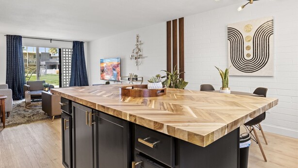A modern kitchen with a large island, black cabinetry, and a wooden countertop. There is a dining area with a table and chairs, a chandelier, and artwork on the walls.