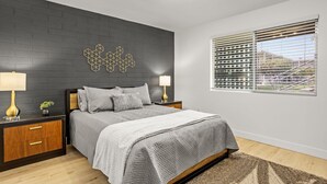 A modern bedroom with a dark brick accent wall, a geometric wall art piece, a large bed with a gray quilt, two bedside lamps, and a window with blinds.