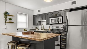 A modern kitchen with dark cabinets, stainless steel appliances, a wooden island with stools, and a window with blinds.
