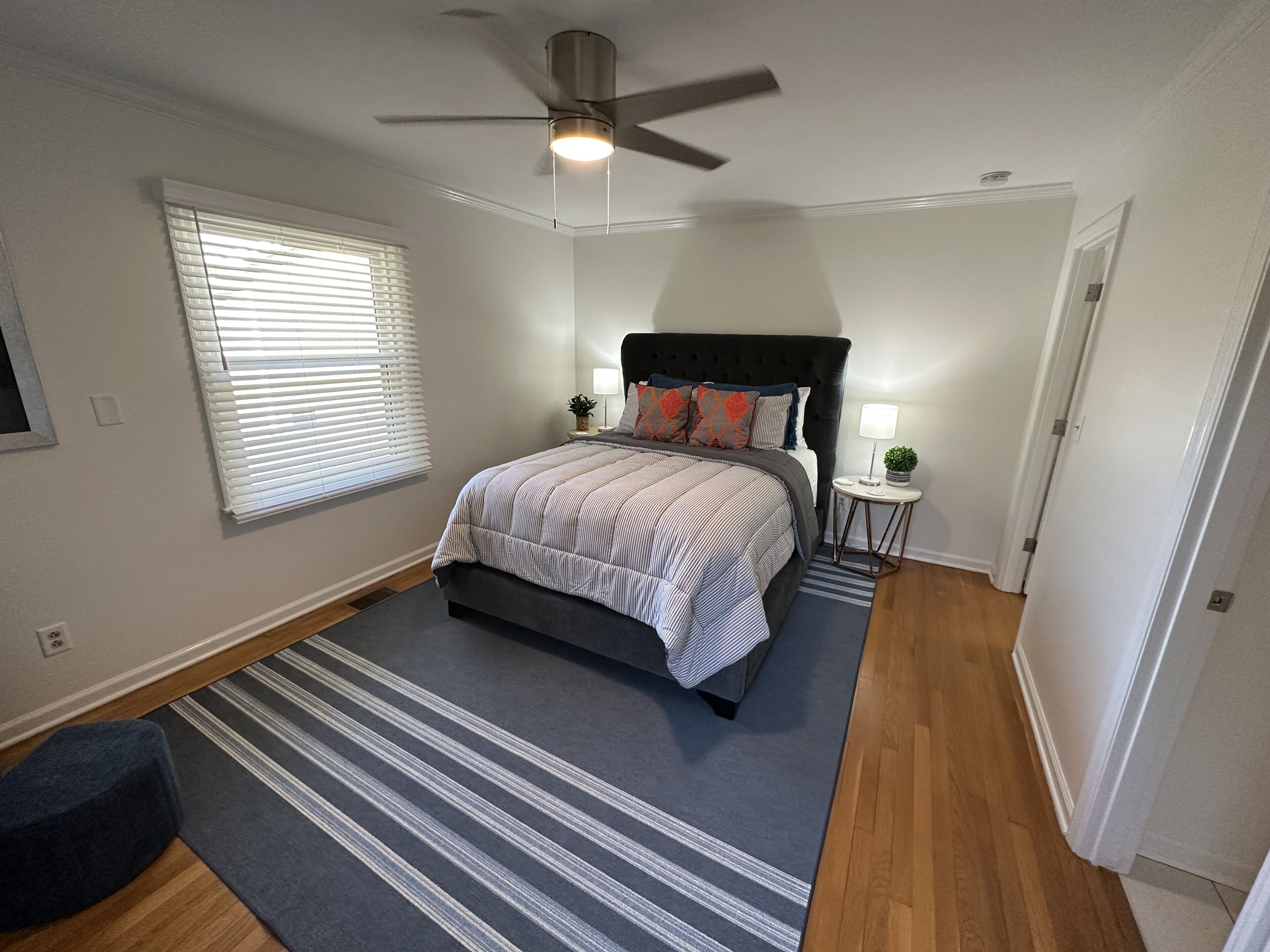 A bedroom with a bed, a ceiling fan, a nightstand with a lamp, and a window with blinds.