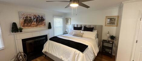 A bedroom with a bed, a ceiling fan, a fireplace, and a painting on the wall.