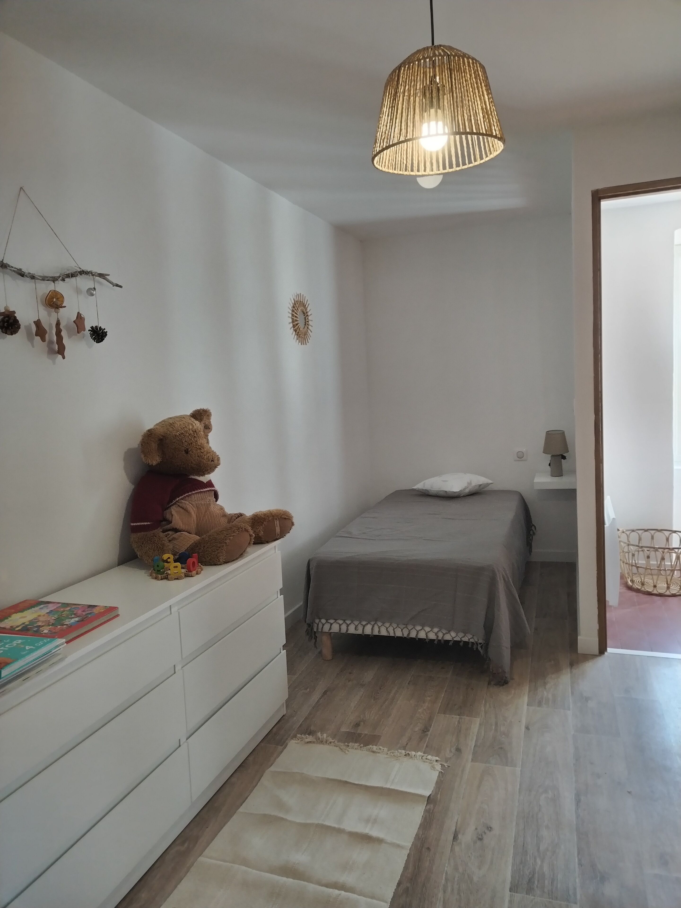 Un dormitorio con cama, cómoda blanca, oso de peluche, lámpara colgante y un pequeño adorno en la pared.