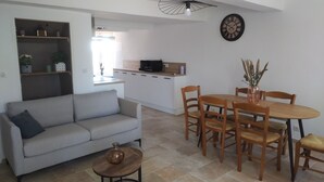 Ein modernes Wohnzimmer mit einer Couch, einem Couchtisch und einem Esstisch mit Stühlen. Es gibt eine Küchenzeile mit einer Mikrowelle und einer Uhr an der Wand.