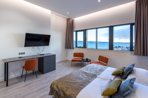 Ein modernes Hotelzimmer mit einem großen Bett, einem Schreibtisch mit Stuhl, einem Fernseher und Blick aufs Meer.