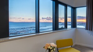 A room with large windows overlooking a coastal area, a yellow chair, and a small table with a vase of flowers. - Seascape Luxury Rooms (Split)