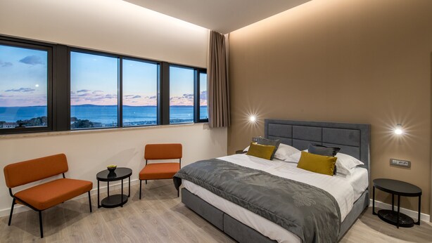 A modern bedroom with a large bed, two chairs, a small table, and a view of the ocean. - Seascape Luxury Rooms (Split)