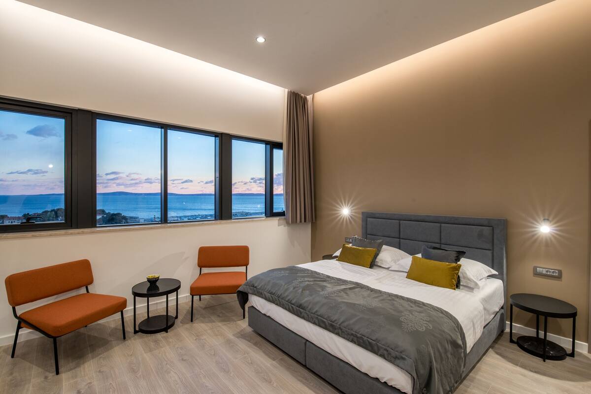 A modern bedroom with a large bed, two chairs, a small table, and a view of the ocean.