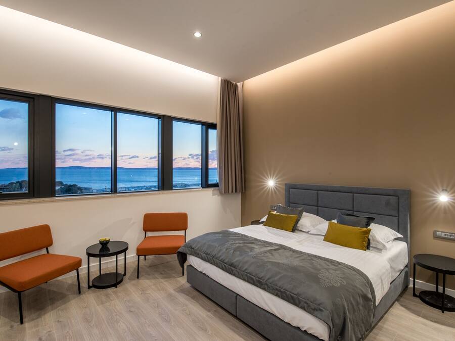 A modern bedroom with a large bed, two chairs, a small table, and a view of the ocean.
