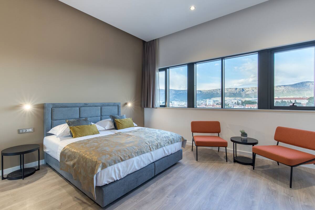 A modern hotel room with a large bed, two chairs, a small table, and a view of a cityscape through large windows.