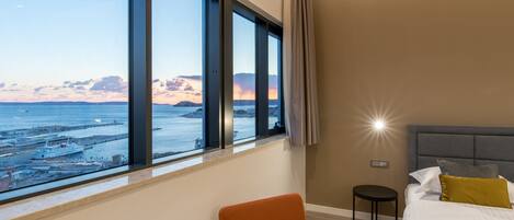 A hotel room with a large window offering a view of the sea and boats, a bed with a grey headboard, and a small round table.