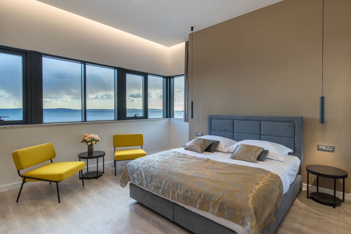 A modern bedroom with a large bed, two yellow chairs, a small table with flowers, and a view of the sea through large windows.