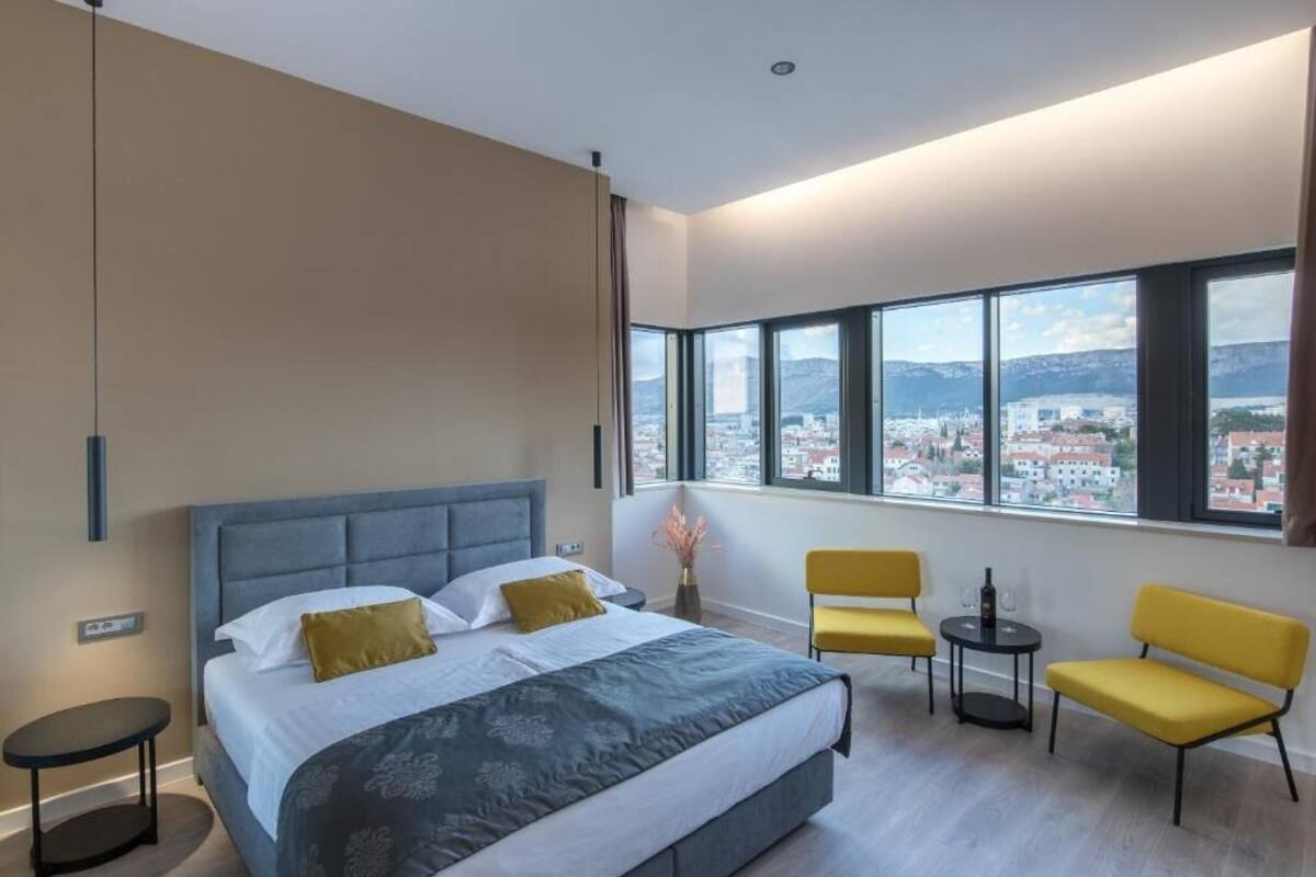 A modern hotel room with a large bed, two yellow chairs, a small table, and a view of a cityscape through large windows.
