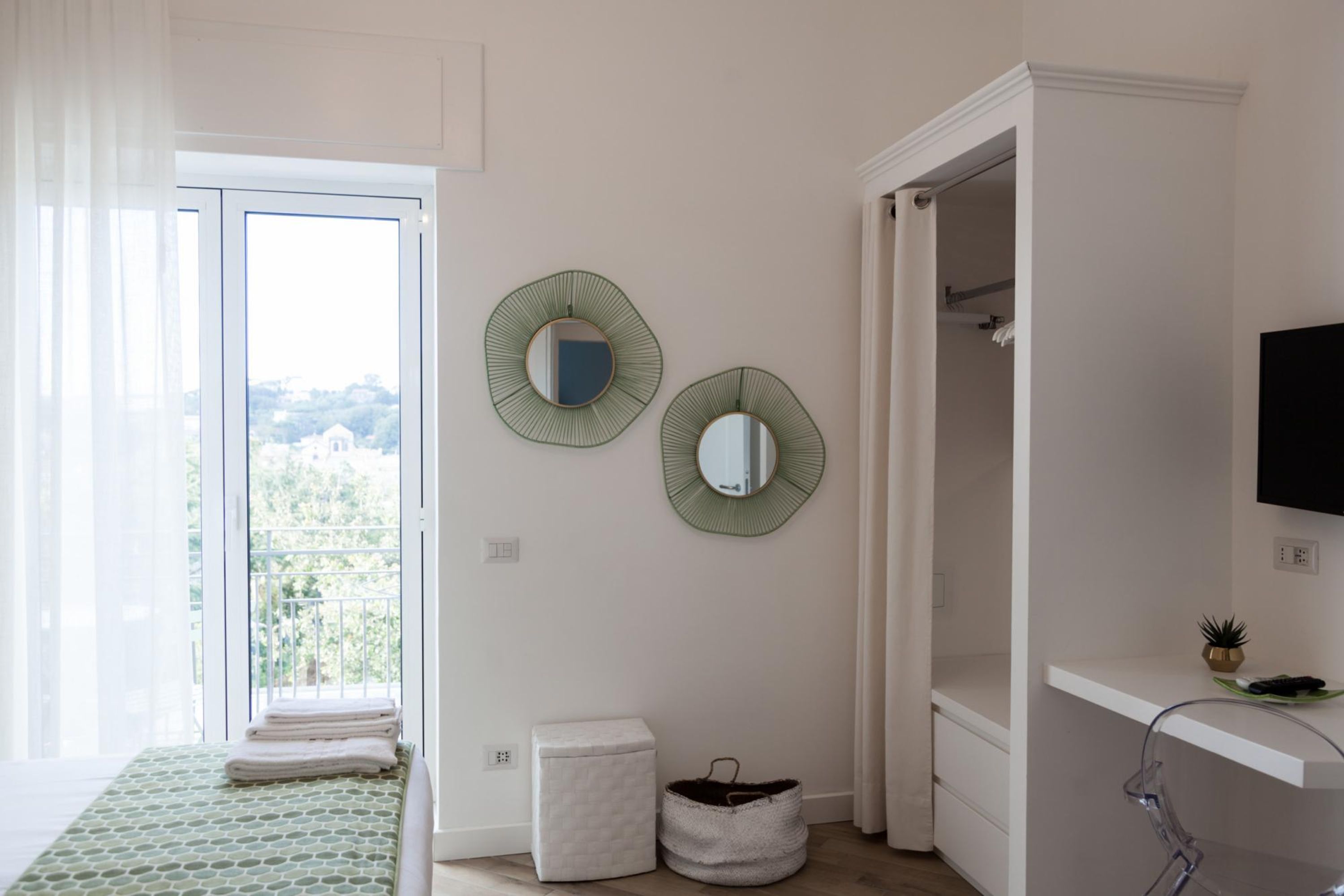 A bedroom with a bed, a small white chest of drawers, a white bench, a round mirror, and a television on a white shelf.