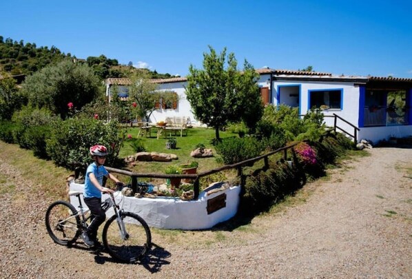 Une personne à vélo, dans un environnement rural, avec un bâtiment blanc, une clôture en bois et un chemin de gravier.