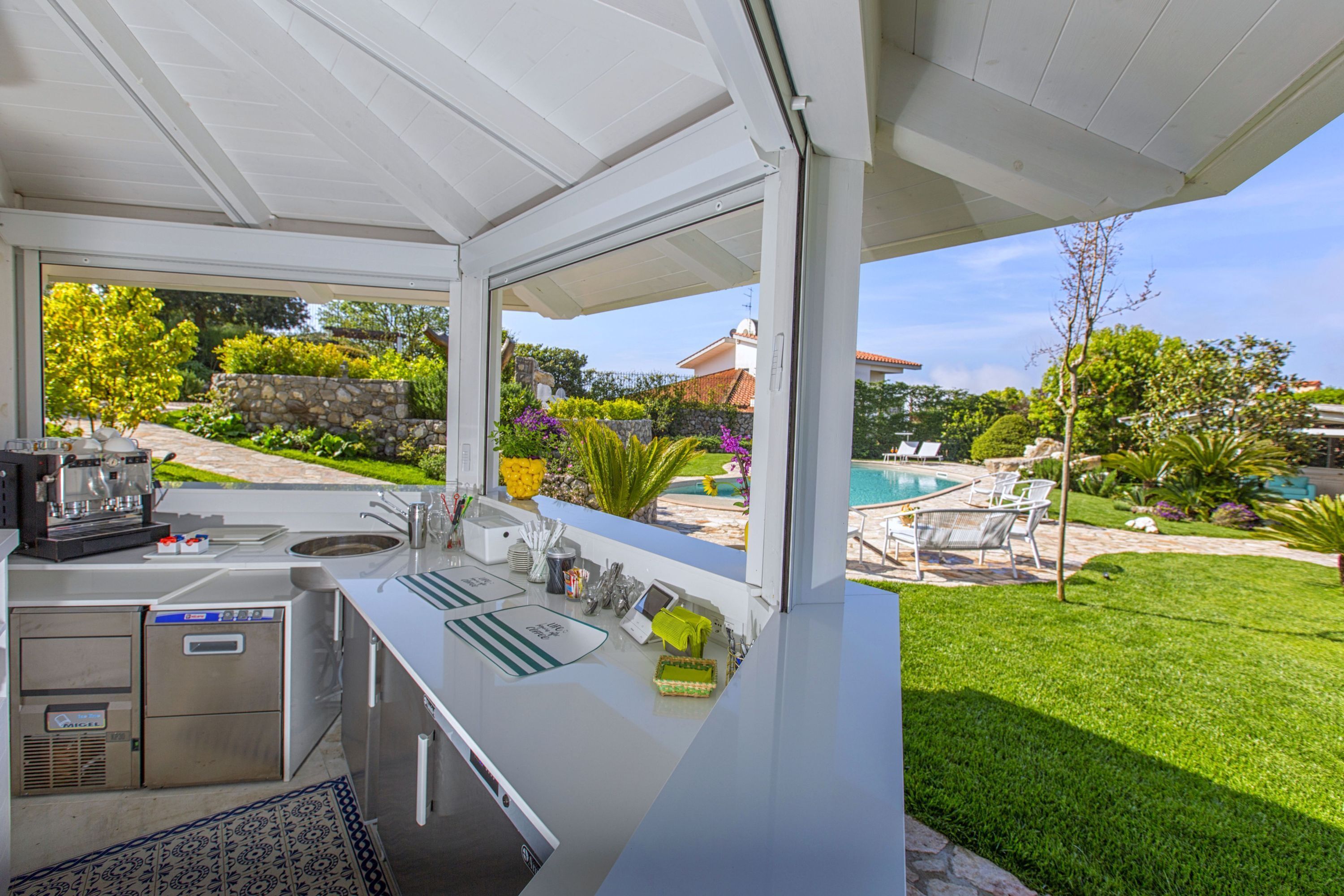 A modern outdoor kitchen with a built-in sink, a coffee machine, and a view of a backyard with a pool and greenery.