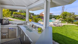 A modern outdoor kitchen with a built-in sink, a coffee machine, and a view of a backyard with a pool and greenery. - Resort Ravenna in Massa Lubrense (Massa Lubrense)