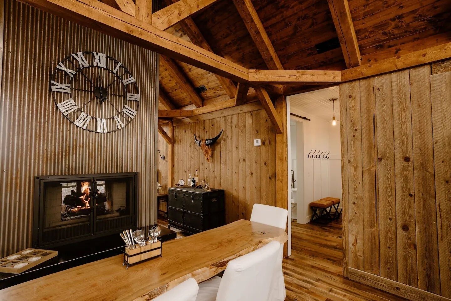 Un intérieur rustique avec un plafond en bois, une grande horloge murale, une cheminée et un coin repas comprenant une table en bois et des chaises blanches.
