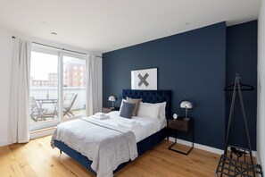 A bedroom with a large bed, a view through a sliding door to an outdoor balcony, and a dark blue accent wall. - home.ly Prestige London Apartment Camden (London)