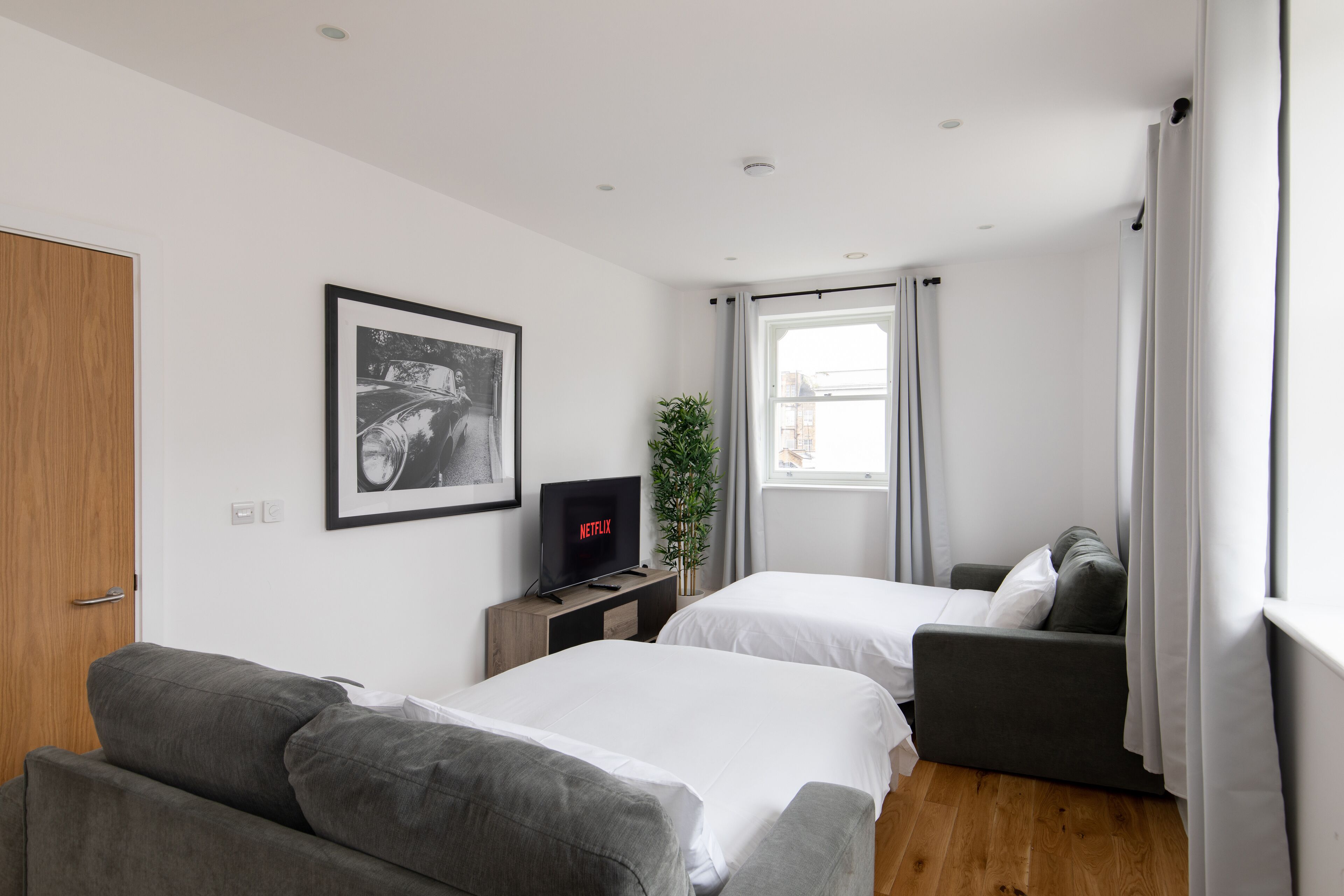 Une chambre d’hôtel moderne avec deux lits, un canapé, une télévision, un tableau encadré et une fenêtre avec des rideaux.