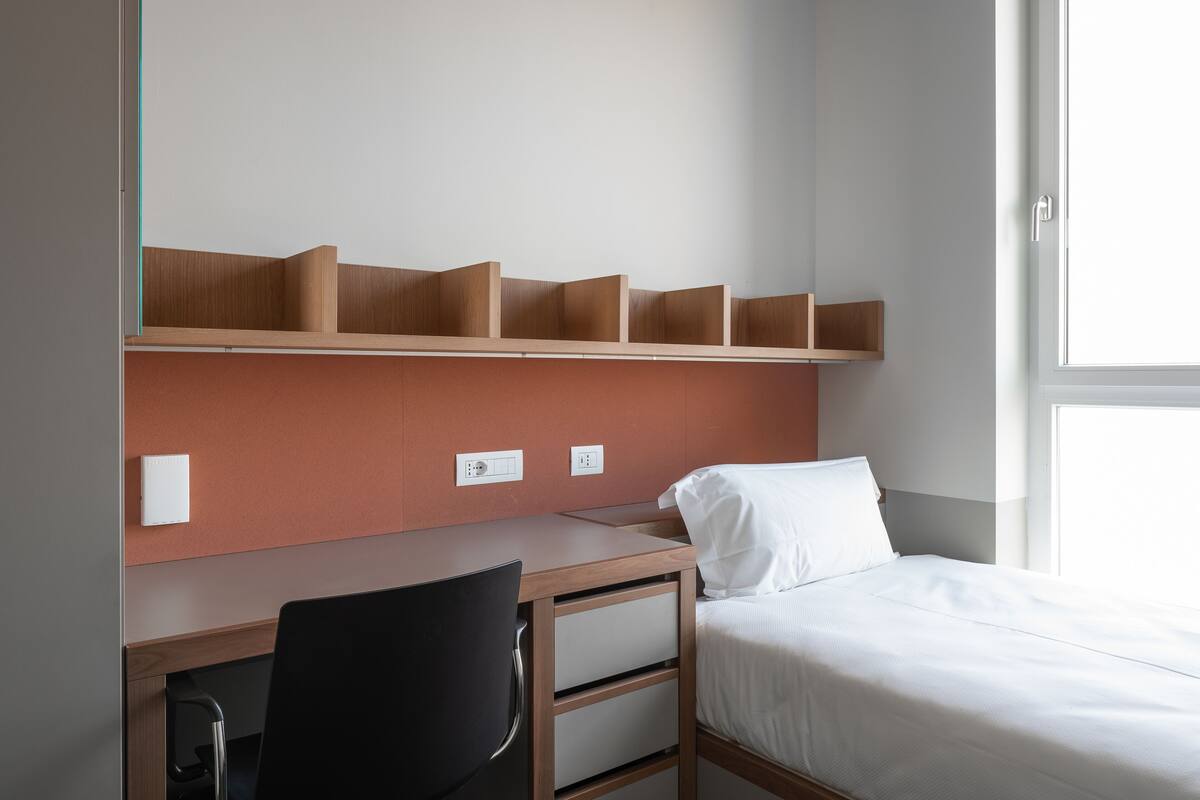 A hotel room with a wooden desk, a black chair, a bed with white bedding, and a window with a view.