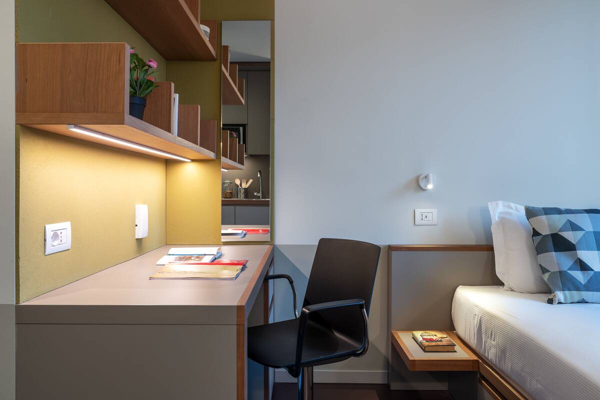 A compact room with a desk, chair, and a bed with a geometric pillow.
