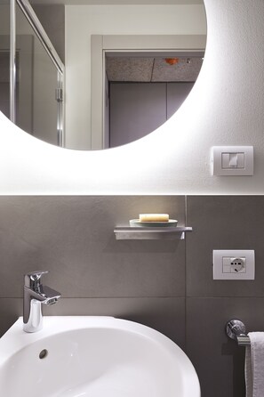 Ein modernes Badezimmer mit einem weißen Waschbecken, einem Chrom-Wasserhahn und einem Spiegel darüber. An der Wand befinden sich ein Handtuchhalter und eine Seifenschale.