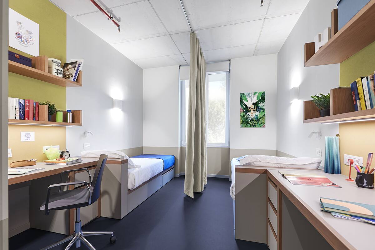A modern hotel room with a desk, two beds, a chair, and shelves with books and decorative items.