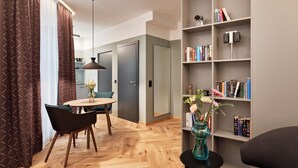 Una sala de estar moderna con piso de madera en patrón de espiga, un comedor con una mesa redonda y sillas, una estantería con libros y adornos, y un ventanal con cortinas.